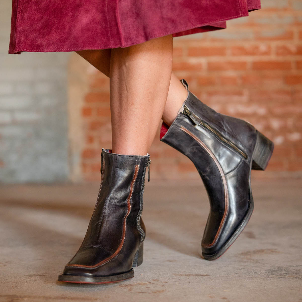 A person in a burgundy skirt wears Bed Stu's Bitsy black ankle boot with a stacked wooden heel, standing on concrete by a brick wall and highlighting the hand-finished leather style.