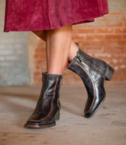 A person in a burgundy skirt wears Bed Stu's Bitsy black ankle boot with a stacked wooden heel, standing on concrete by a brick wall and highlighting the hand-finished leather style.