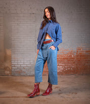 A woman stands indoors against a brick wall, wearing wide-leg jeans, a blue striped shirt, a brown belt, and red Bed Stu Bitsy ankle boots with a stacked wooden heel.