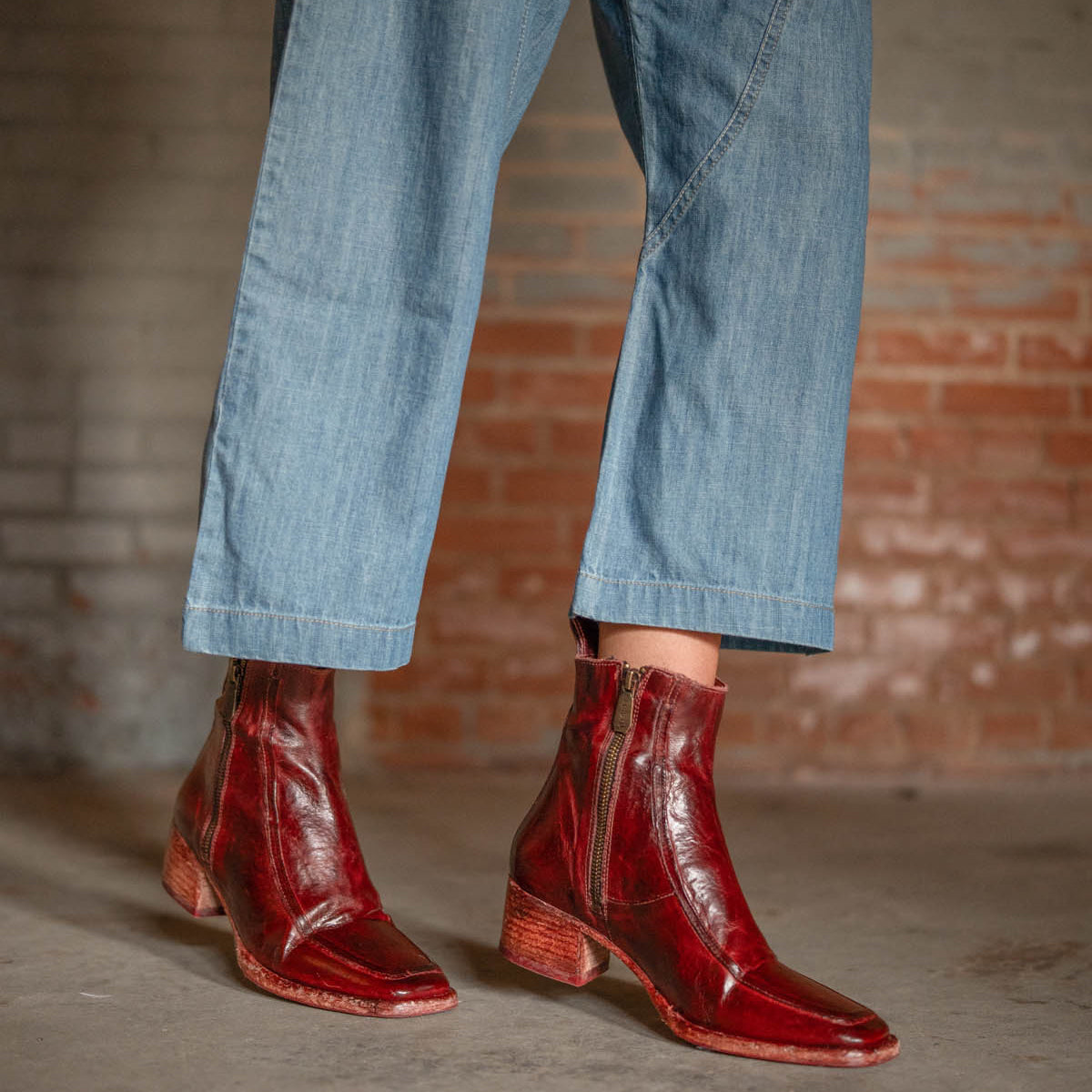 A person stands on a concrete floor by a brick wall, wearing cropped blue jeans and Bed Stu's Bitsy—shiny, hand-finished red leather ankle boots with side zippers and stacked wooden heels.