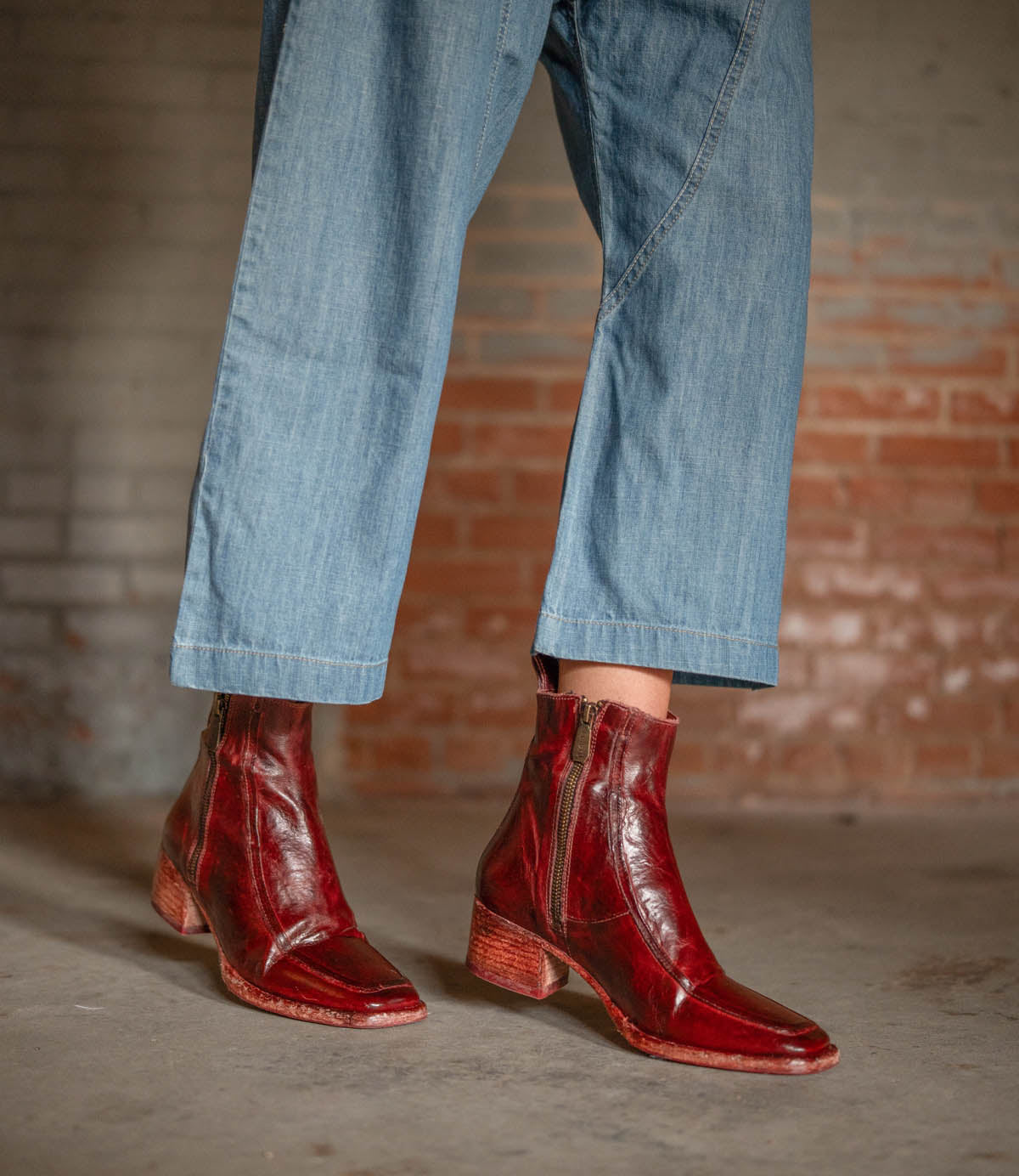 A person stands on a concrete floor by a brick wall, wearing cropped blue jeans and Bed Stu's Bitsy—shiny, hand-finished red leather ankle boots with side zippers and stacked wooden heels.