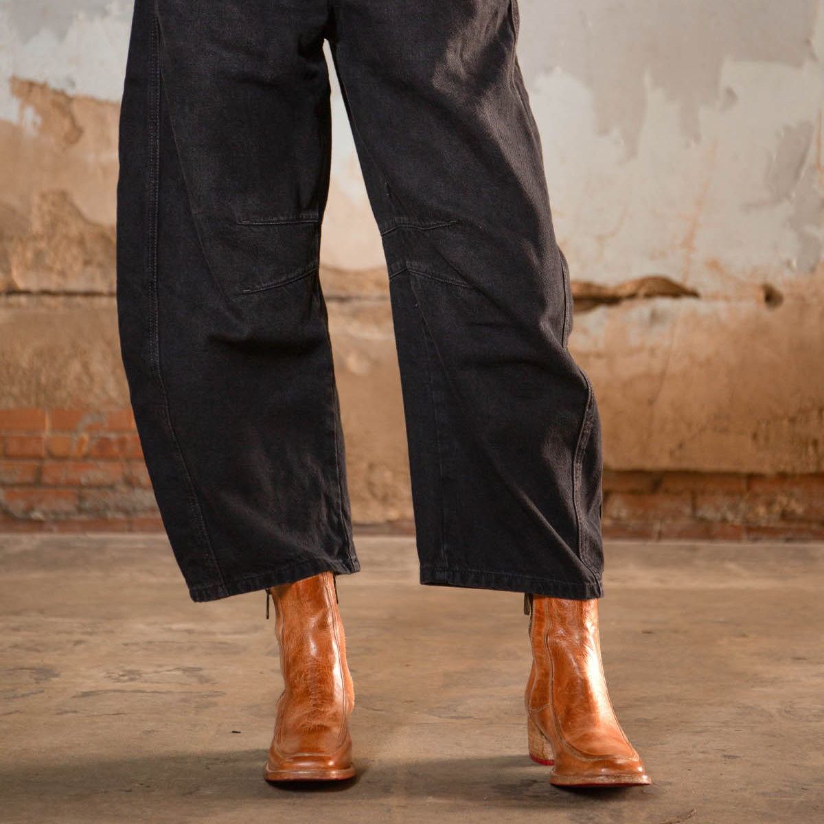 A person wearing loose black pants and Bed Stu's Bitsy brown hand-finished leather ankle boots with a stacked wooden heel stands on a concrete floor against a weathered, textured wall.