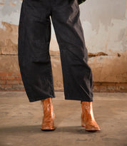 A person wearing loose black pants and Bed Stu's Bitsy brown hand-finished leather ankle boots with a stacked wooden heel stands on a concrete floor against a weathered, textured wall.