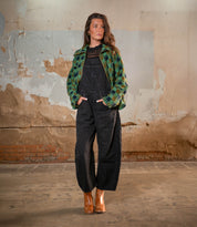 A woman stands indoors against a distressed wall, hands in pockets, wearing black overalls, a green checkered jacket, and Bed Stu Bitsy brown stacked wooden heel ankle boots, gazing to the side.