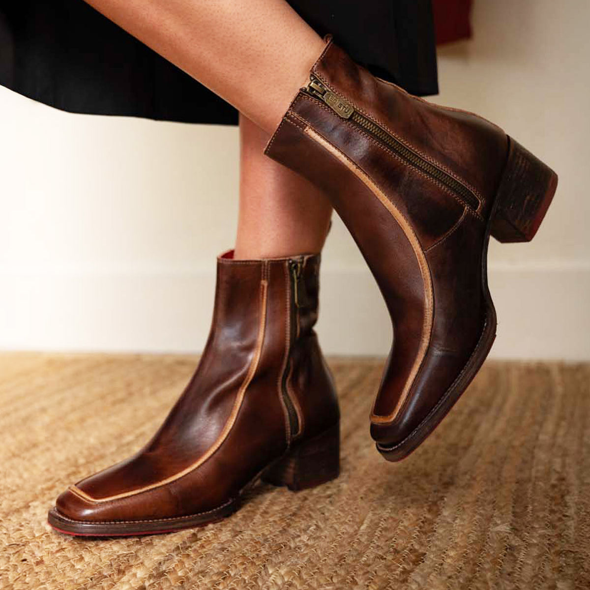 A person wearing the Bed|Stü Bitsy mid-calf brown leather boots, featuring visible zippers and a stacked heel, stands on a woven rug.