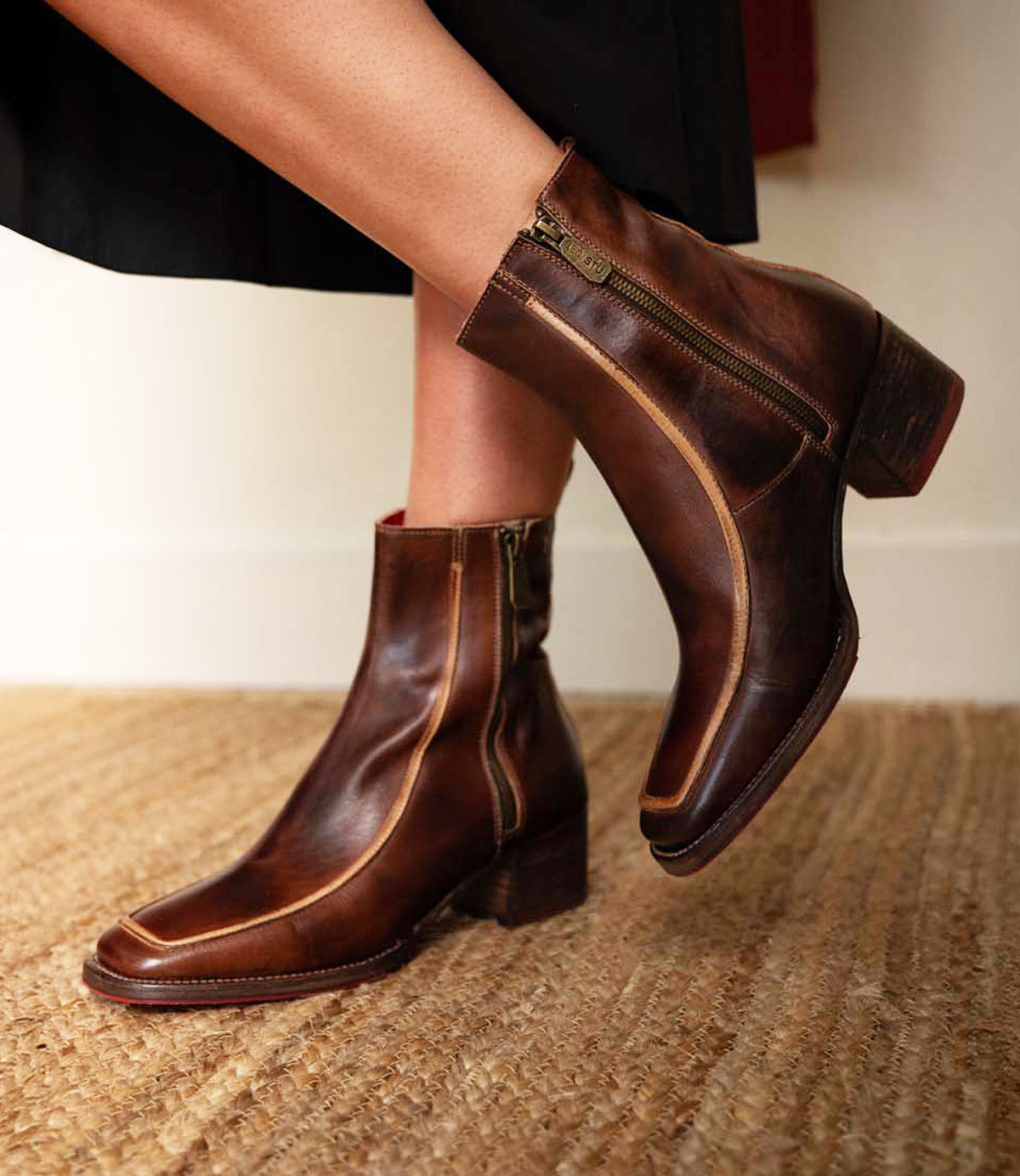 A person wearing the Bed|Stü Bitsy mid-calf brown leather boots, featuring visible zippers and a stacked heel, stands on a woven rug.