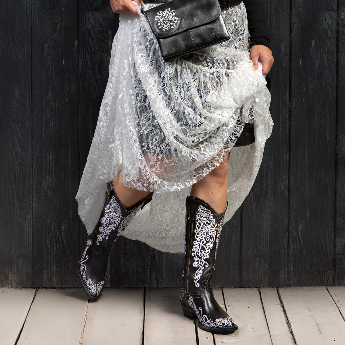Wearing the Bed Stu Blackjack white lace skirt and Western tall boots with floral embroidery, holding a black purse with white floral design, standing on wooden floor against a dark wall.