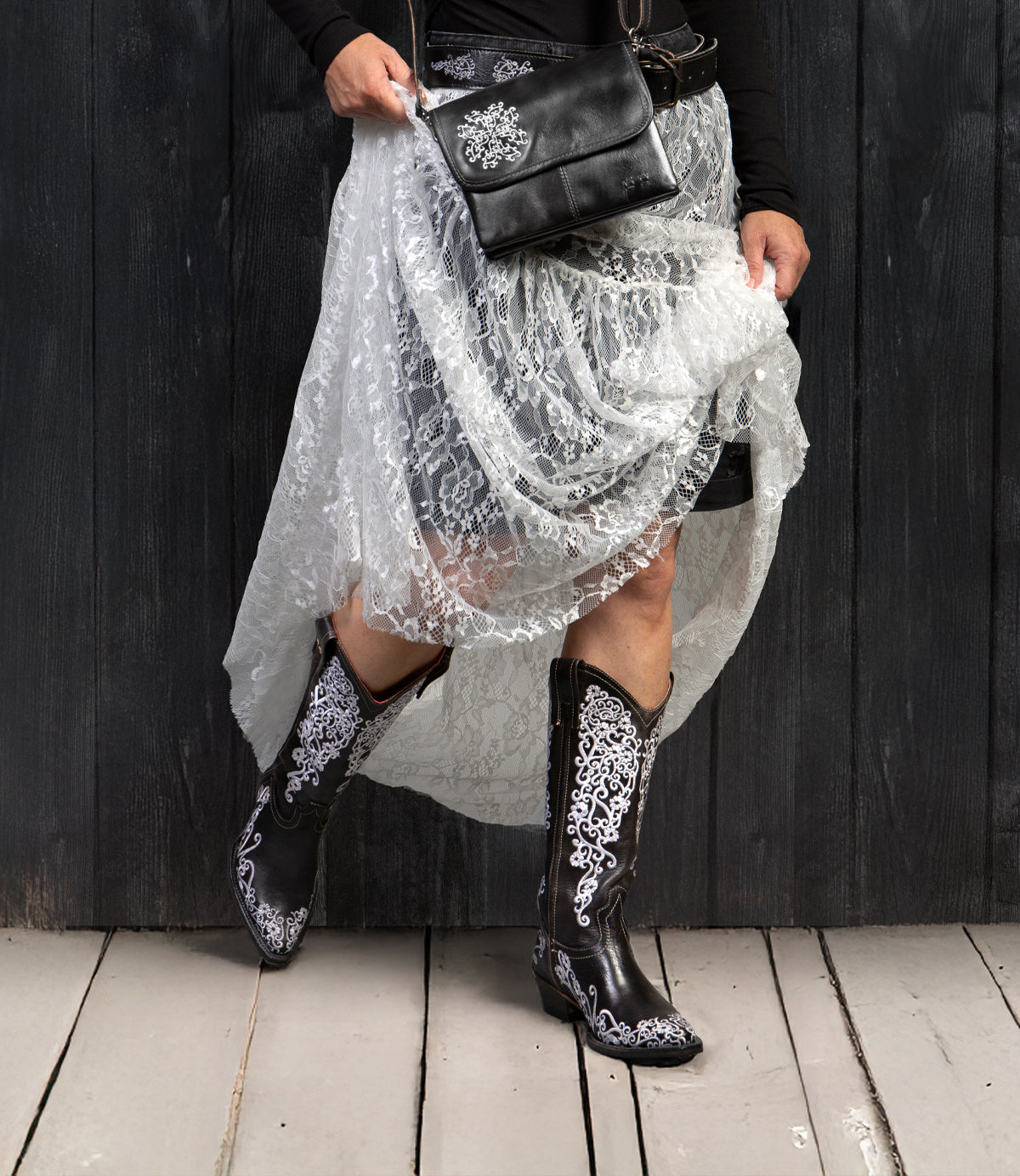 Wearing the Bed Stu Blackjack white lace skirt and Western tall boots with floral embroidery, holding a black purse with white floral design, standing on wooden floor against a dark wall.