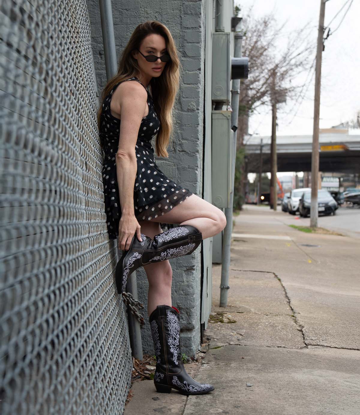 A woman in a black polka dot dress and Bed Stu Blackjack Western tall boots with floral embroidery leans against a city chain-link fence, looking at the camera.