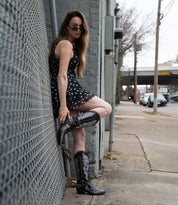 A woman in a black polka dot dress and Bed Stu Blackjack Western tall boots with floral embroidery leans against a city chain-link fence, looking at the camera.