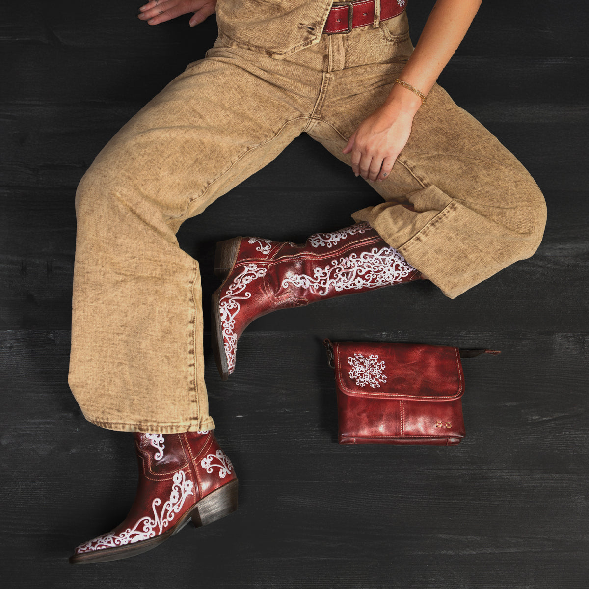 Wearing Bed Stu Blackjack tan overalls and Western tall boots with red floral embroidery, a person sits on a dark surface next to a matching red leather bag.