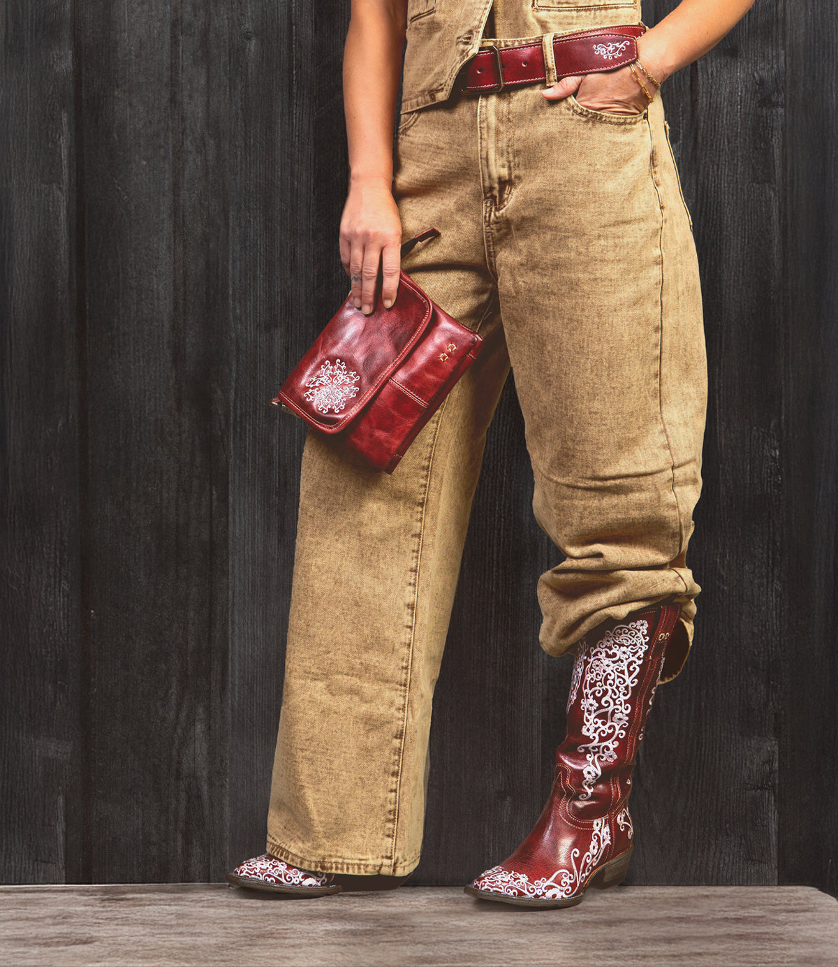 A person stands against a dark wooden wall, wearing tan jeans, a red belt, holding a matching red clutch, and sporting the Bed Stu Blackjack Western tall boots with white embroidery.