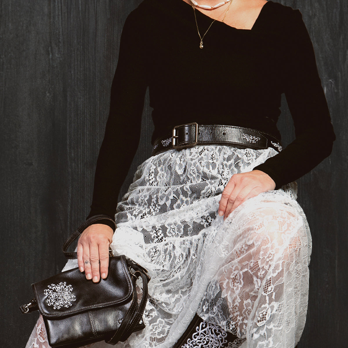 A woman sits against a dark background wearing a black top, white lace skirt, and a handcrafted belt, holding the Bed Stu Blaze JB black purse with embroidered scrollwork.