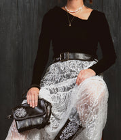 A woman sits against a dark background wearing a black top, white lace skirt, and a handcrafted belt, holding the Bed Stu Blaze JB black purse with embroidered scrollwork.