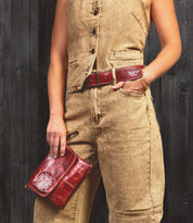 A person models the Bed Stu Blaze JB tan denim jumpsuit with one hand in a pocket, holding a red leather clutch, and wearing a handcrafted belt featuring white decorative details.