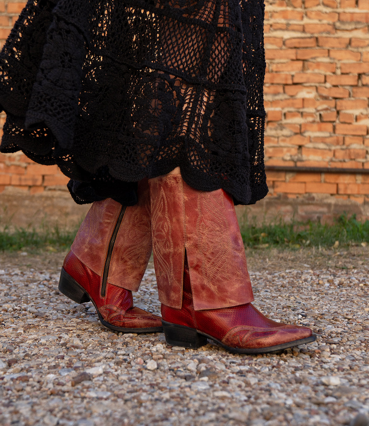 A person wearing the Blind long black crocheted skirt and Bed Stu red distressed leather Western boots walks on a gravel path with a brick wall in the background.