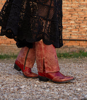A person wearing the Blind long black crocheted skirt and Bed Stu red distressed leather Western boots walks on a gravel path with a brick wall in the background.