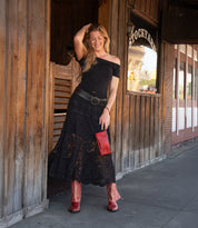 A woman wears Bed Stu's Blind red Western boots, holding a matching clutch and smiling in a black off-shoulder top and skirt near a wooden Stockyard building.