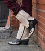 A person wearing Bed Stu’s Blind boots—light-colored, patterned cowboy boots with a Western silhouette and dark toes and heels—leans against a brick wall, one foot crossed over the other.