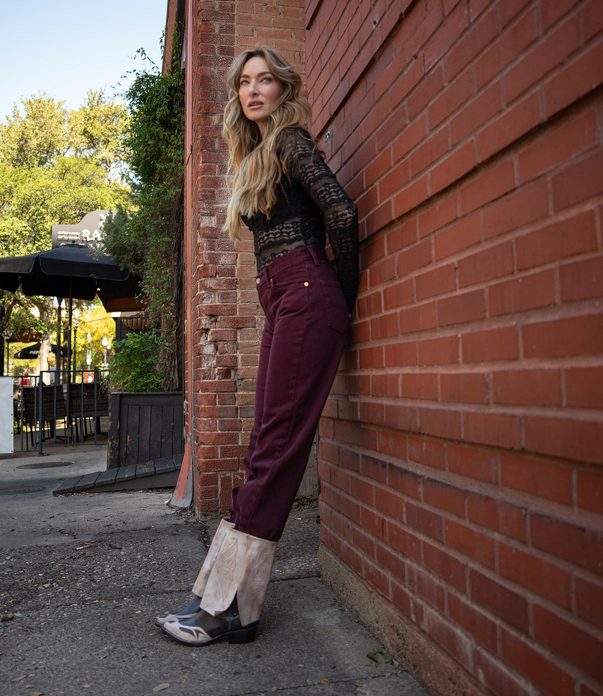 A woman with long hair stands by a red brick wall, wearing a black lace top, maroon pants with tan cuffs, and Bed Stu's Blind foldover boots, which bring a Western-inspired touch to her outfit.