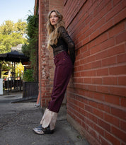 A woman with long hair stands by a red brick wall, wearing a black lace top, maroon pants with tan cuffs, and Bed Stu's Blind foldover boots, which bring a Western-inspired touch to her outfit.