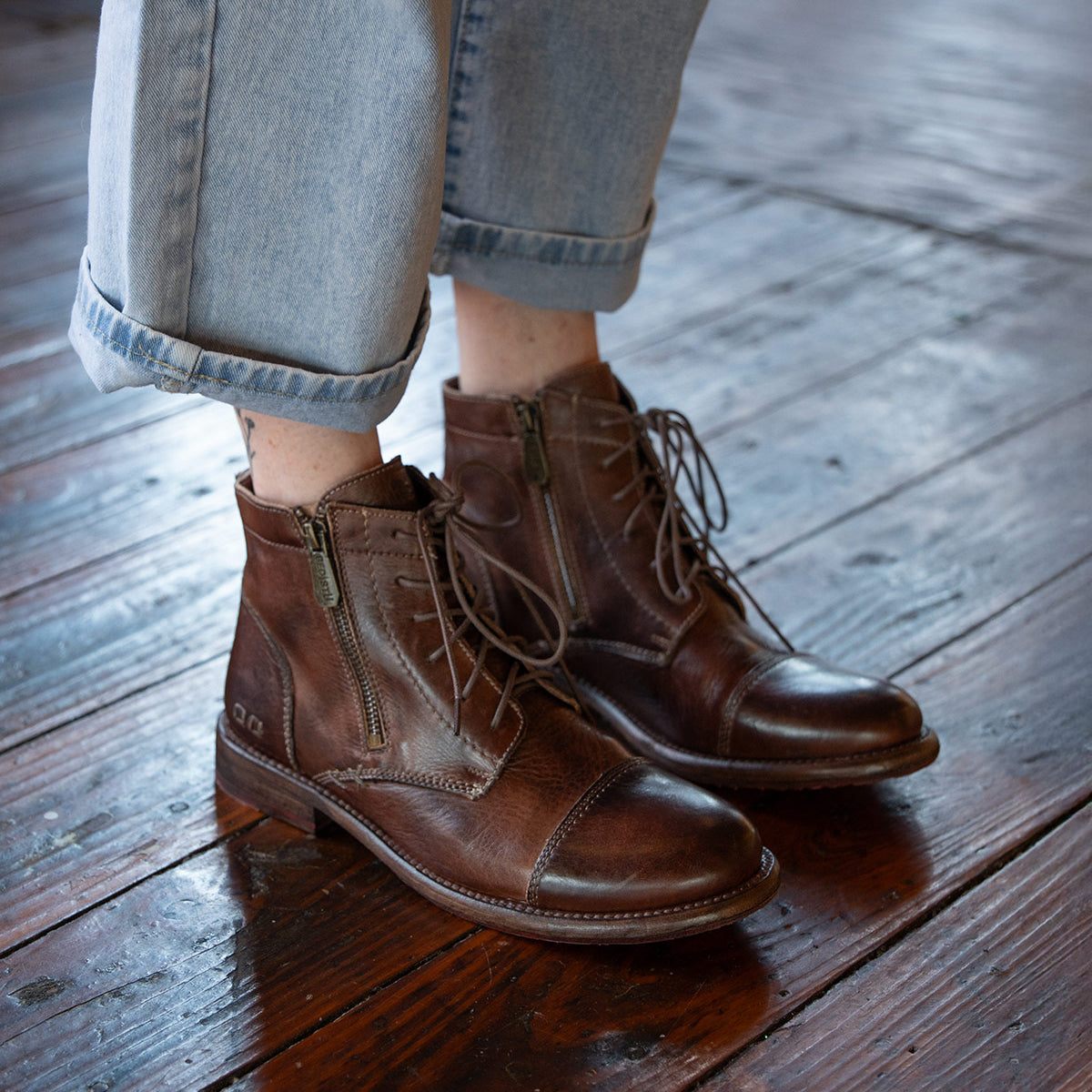 A person wearing Bed Stu Bonnie II brown lace-up Goodyear-welted ankle boots with a cap toe and light blue cuffed jeans stands on a polished wooden floor.