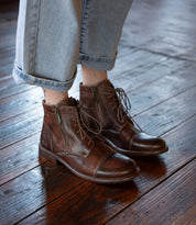 A person wearing Bed Stu Bonnie II brown lace-up Goodyear-welted ankle boots with a cap toe and light blue cuffed jeans stands on a polished wooden floor.