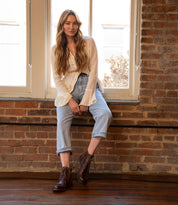 A woman with long hair sits on a windowsill in front of a brick wall, wearing a white shirt, light blue jeans, and Bed Stu’s Bonnie II cap toe combat ankle boots.