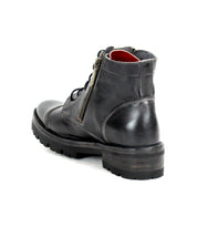 The Bed|Stü Bonnie IV is a black leather combat boot with a no-frills silhouette, visible stitching, lace-up front, dual side zippers, and a thick rubber sole, shown from the back angle on a white background.