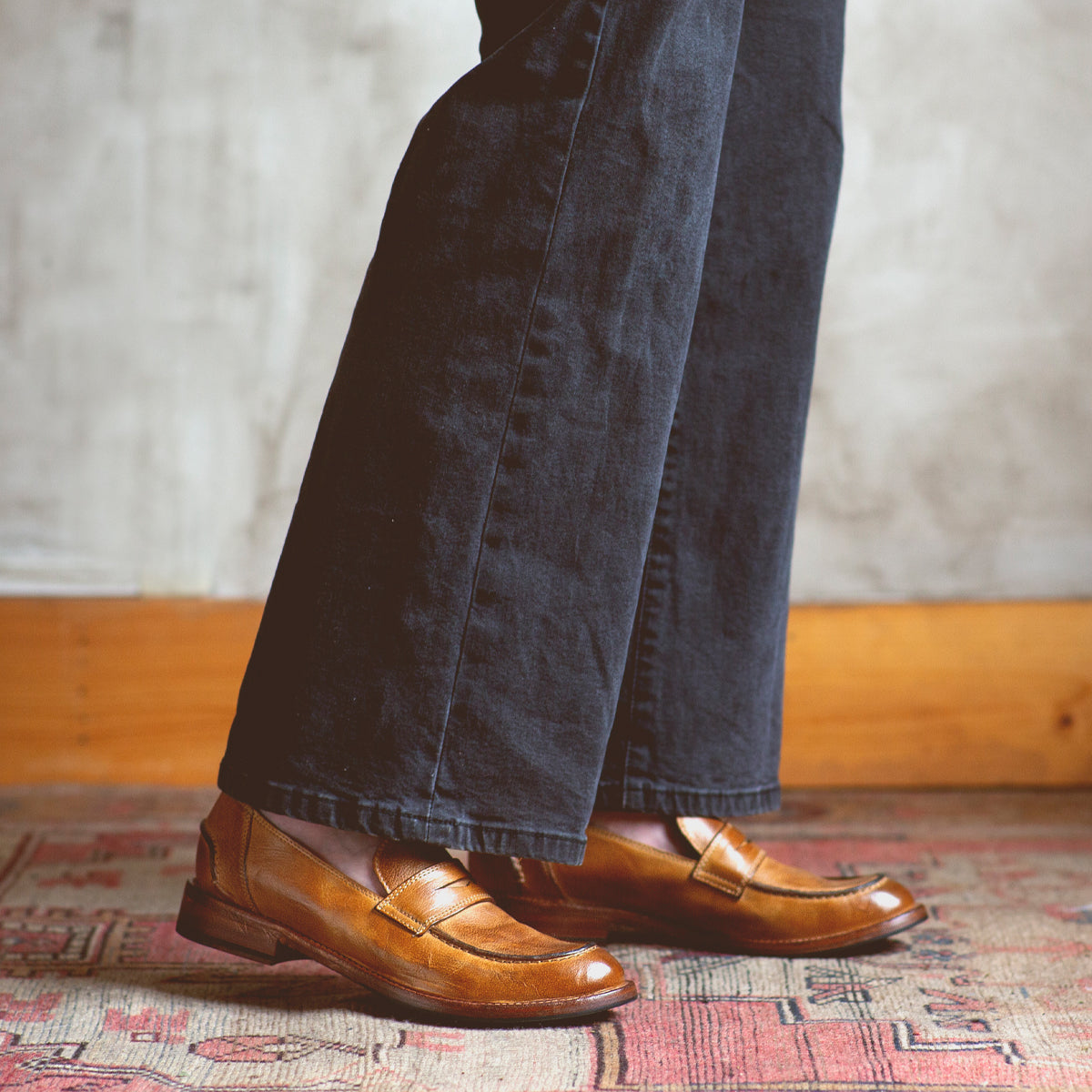 A person standing on a rug wearing a pair of Bed Stu Bonus penny loafers crafted from Italian leather.