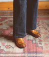 A person standing on a rug wearing a pair of Bed Stu Bonus Italian leather brown shoes with a distressed finish.