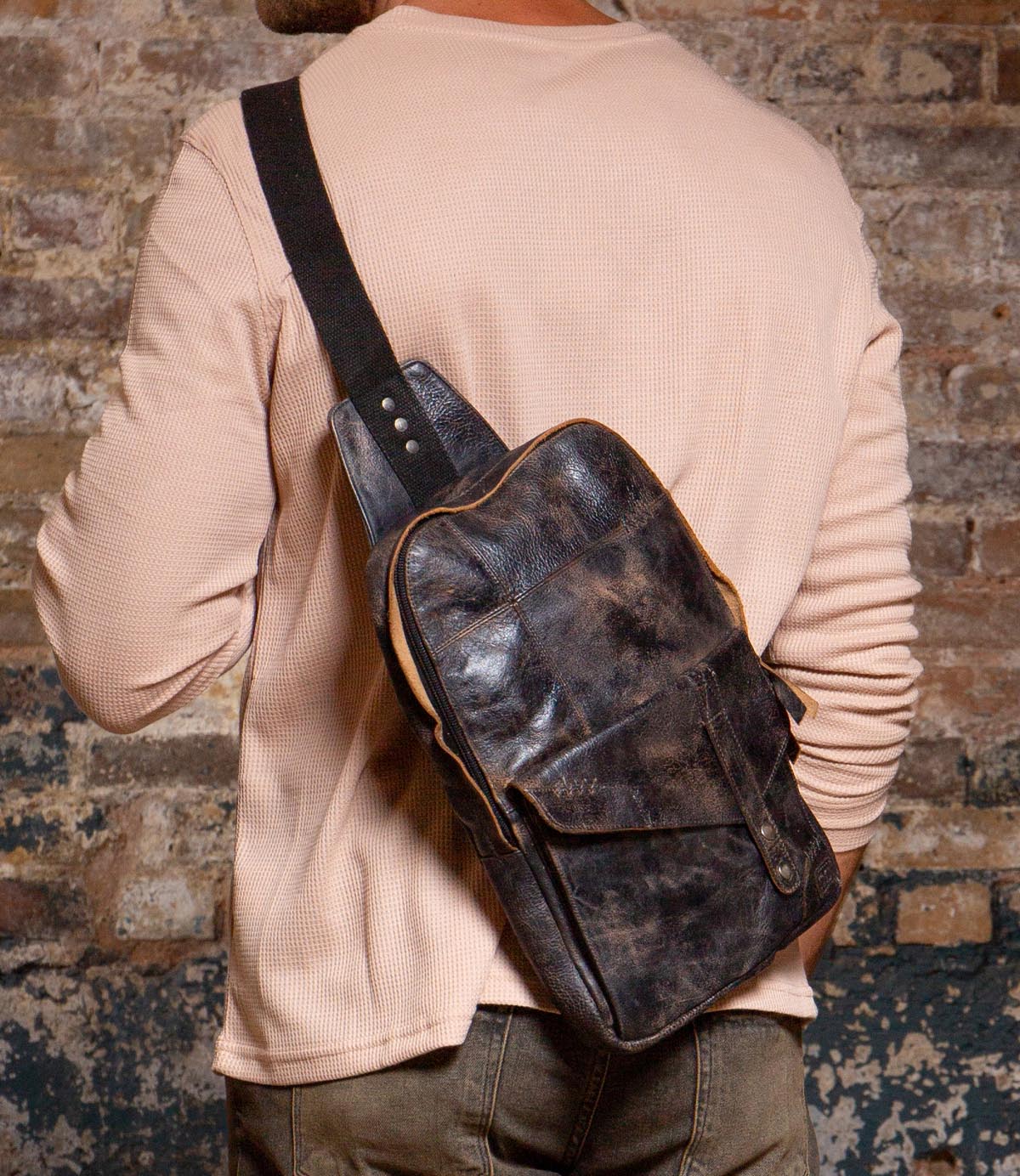 A person in a beige long-sleeve shirt and jeans carries the Bed Stu Boss dark brown leather sling bag with a tablet compartment over their shoulder, standing before a textured brick wall.