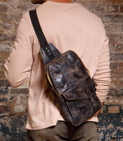 A person in a beige long-sleeve shirt and jeans carries the Bed Stu Boss dark brown leather sling bag with a tablet compartment over their shoulder, standing before a textured brick wall.
