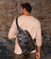 A man in a beige long-sleeve shirt stands before a textured brick wall, carrying the Bed Stu Boss—a durable dark leather sling bag with a tablet compartment.