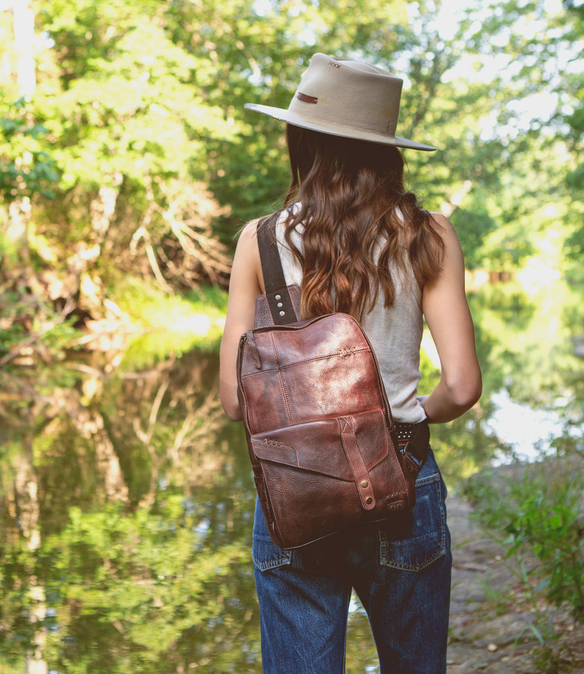 A woman wearing a hat and jeans standing by a river with a Bed Stu leather sling backpack named Beau.