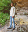 A man in a white shirt, jeans, and Bed Stu Black Rustic Boots stands on a rocky path under a cliff covered with green foliage.