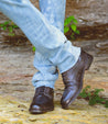 Person wearing rolled-up blue jeans and Bed Stu Black Rustic Boots standing on a yellow painted curb.