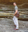 A man in a striped shirt and white pants, sporting rustic boots, carries a Bed Stu brown leather bag and stands on a rocky ledge, looking over his shoulder.