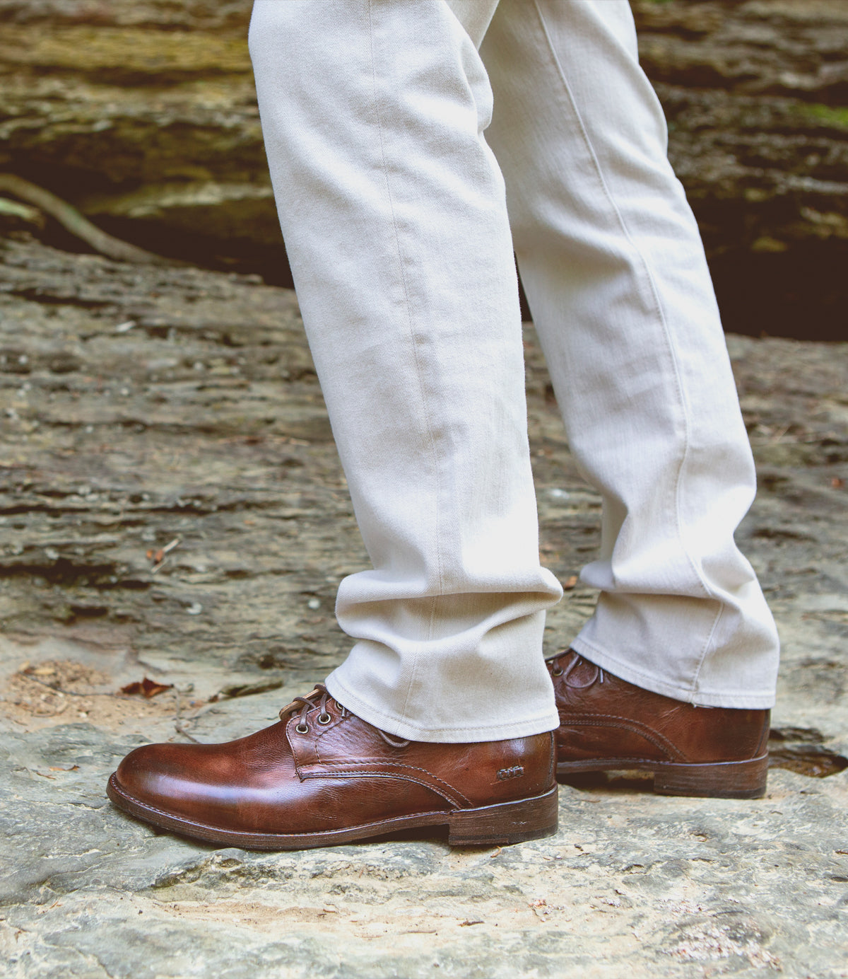 A close-up view of a person wearing beige trousers and brown Bed Stu Black Rustic Boots standing on rocky terrain.