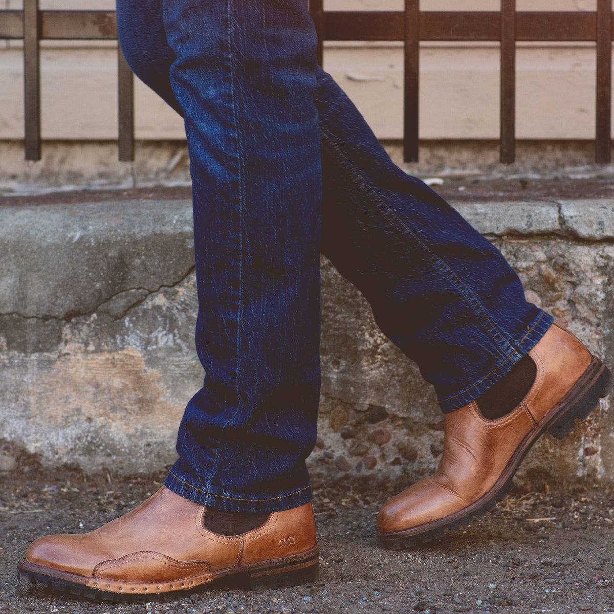Close-up of a person wearing blue jeans and the Brady Trek brown leather Chelsea boots with a Vibram outsole by Bed Stu, standing on a concrete pathway in front of a metal railing.