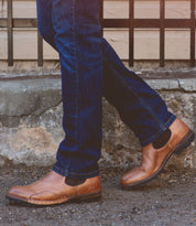 Close-up of a person wearing blue jeans and the Brady Trek brown leather Chelsea boots with a Vibram outsole by Bed Stu, standing on a concrete pathway in front of a metal railing.