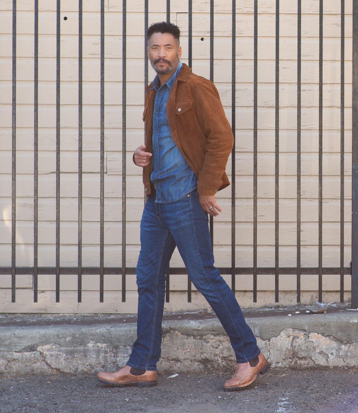 A man wearing a brown suede jacket, a blue denim shirt, blue jeans, and Bed Stu's Brady Trek brown Chelsea boots with a Vibram outsole walks confidently past a background with black vertical bars and a light-colored wall.