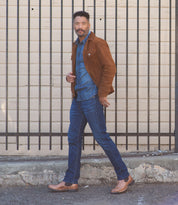 A man wearing a brown suede jacket, a blue denim shirt, blue jeans, and Bed Stu's Brady Trek brown Chelsea boots with a Vibram outsole walks confidently past a background with black vertical bars and a light-colored wall.