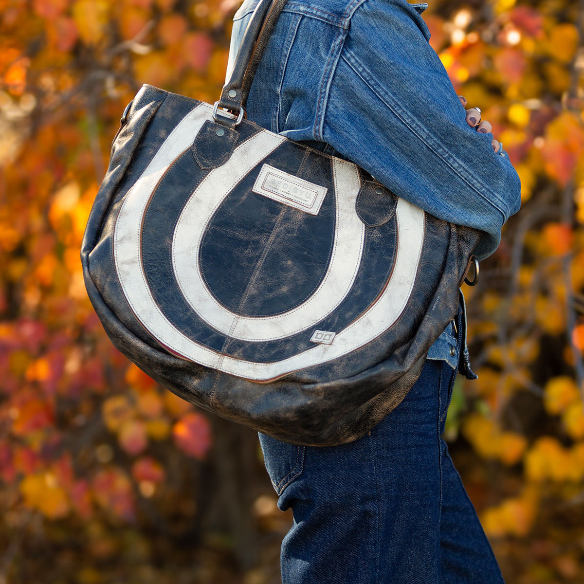 A person wearing a denim jacket and jeans stands against a backdrop of autumn leaves, holding the Bed Stu Brawler carryall that features a striking black and white circular pattern.