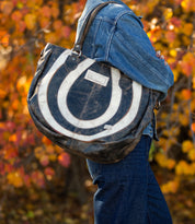 A person wearing a denim jacket and jeans stands against a backdrop of autumn leaves, holding the Bed Stu Brawler carryall that features a striking black and white circular pattern.
