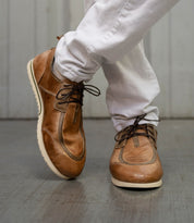 A person wearing white pants and refined brown leather Bed|Stü Britches chukka-style lace-up shoes stands on a wooden floor with one foot crossed over the other.