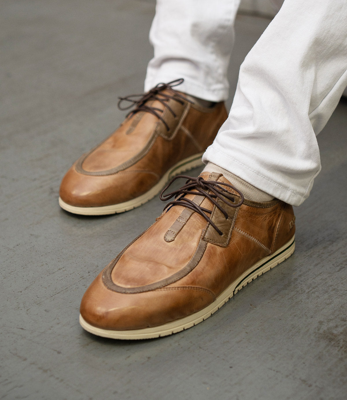 A person wearing the Bed|Stü Britches refined light brown chukka-style leather sneakers with white soles and white pants stands on a gray floor.