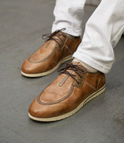 A person wearing the Bed|Stü Britches refined light brown chukka-style leather sneakers with white soles and white pants stands on a gray floor.