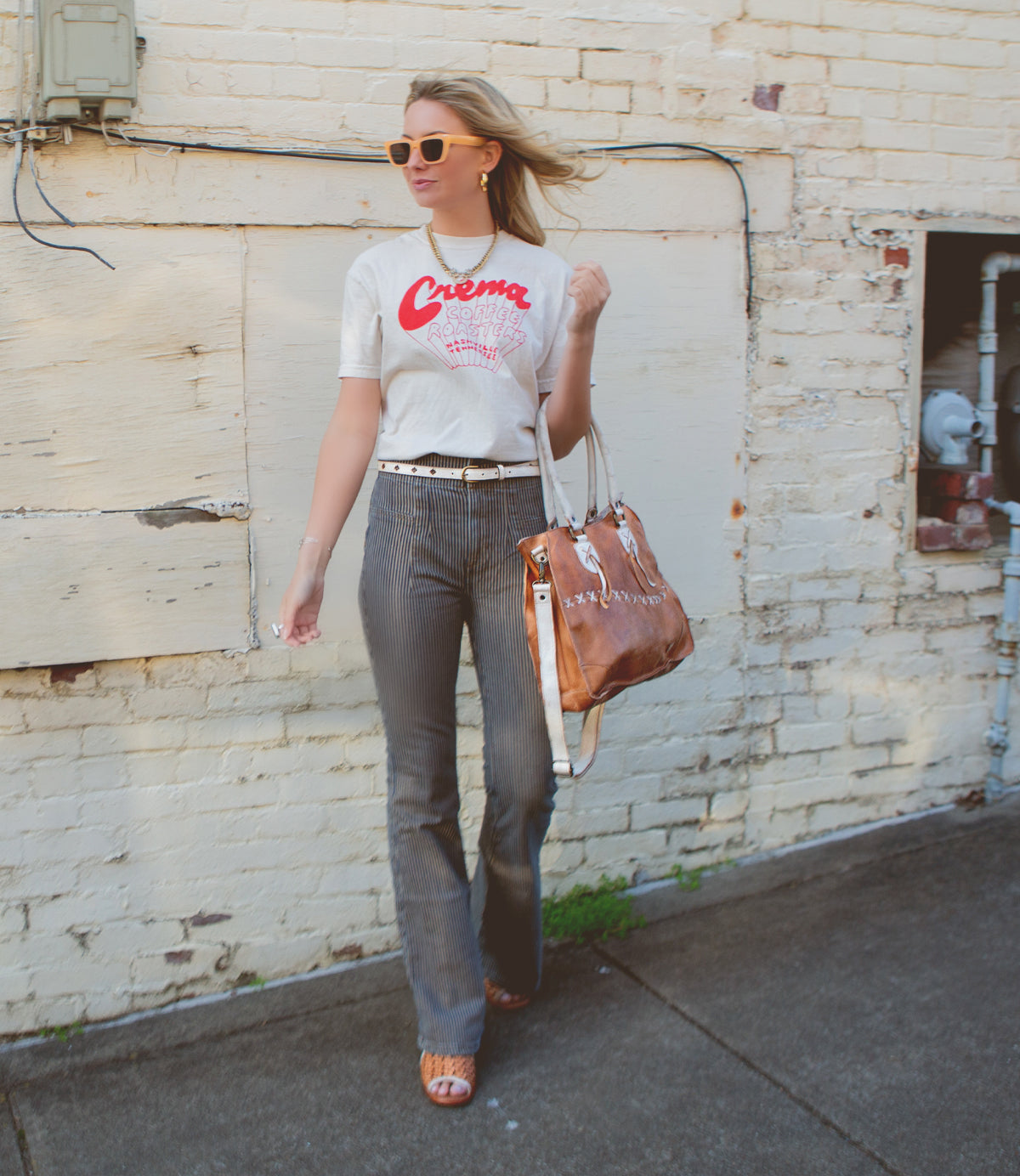 A woman in sunglasses wearing a white t-shirt and striped trousers carries a Bed Stu Bruna two-toned leather bag with shoulder straps while walking past a white brick wall.