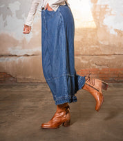 Wearing Bed Stu’s Cable hand-finished leather mid-ankle boots, this person pairs them with loose denim pants, standing indoors against a textured wall for a subtle Western vibe.